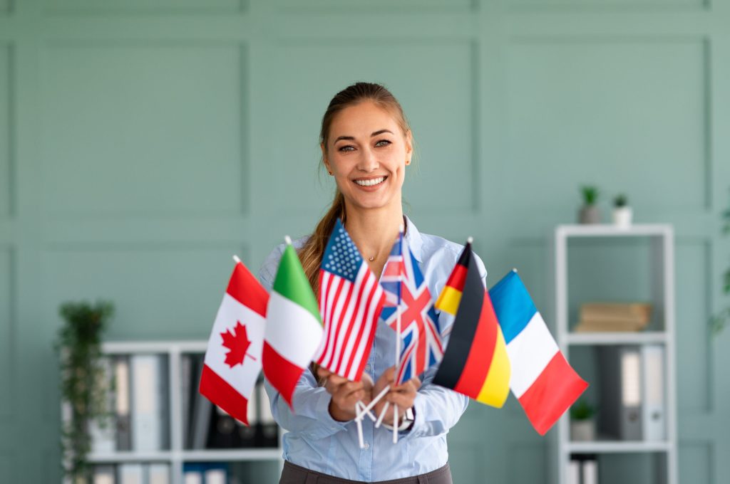 Happy female tutor showing bunch of diverse flags cheerfully smiling at camera, friendly teacher recommending foreign language studying school, posing in office